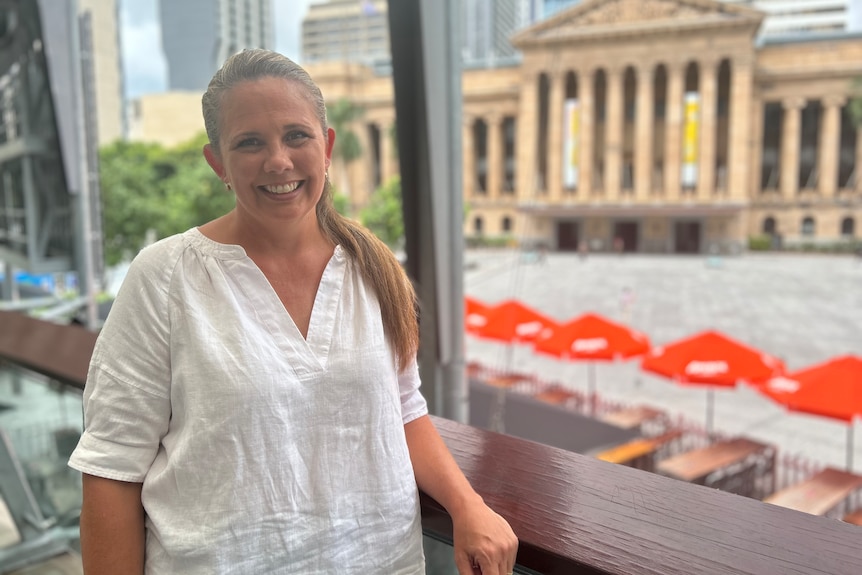 A smiling woman wearing a white shirt standing above a public square with a sandstone building behind her.