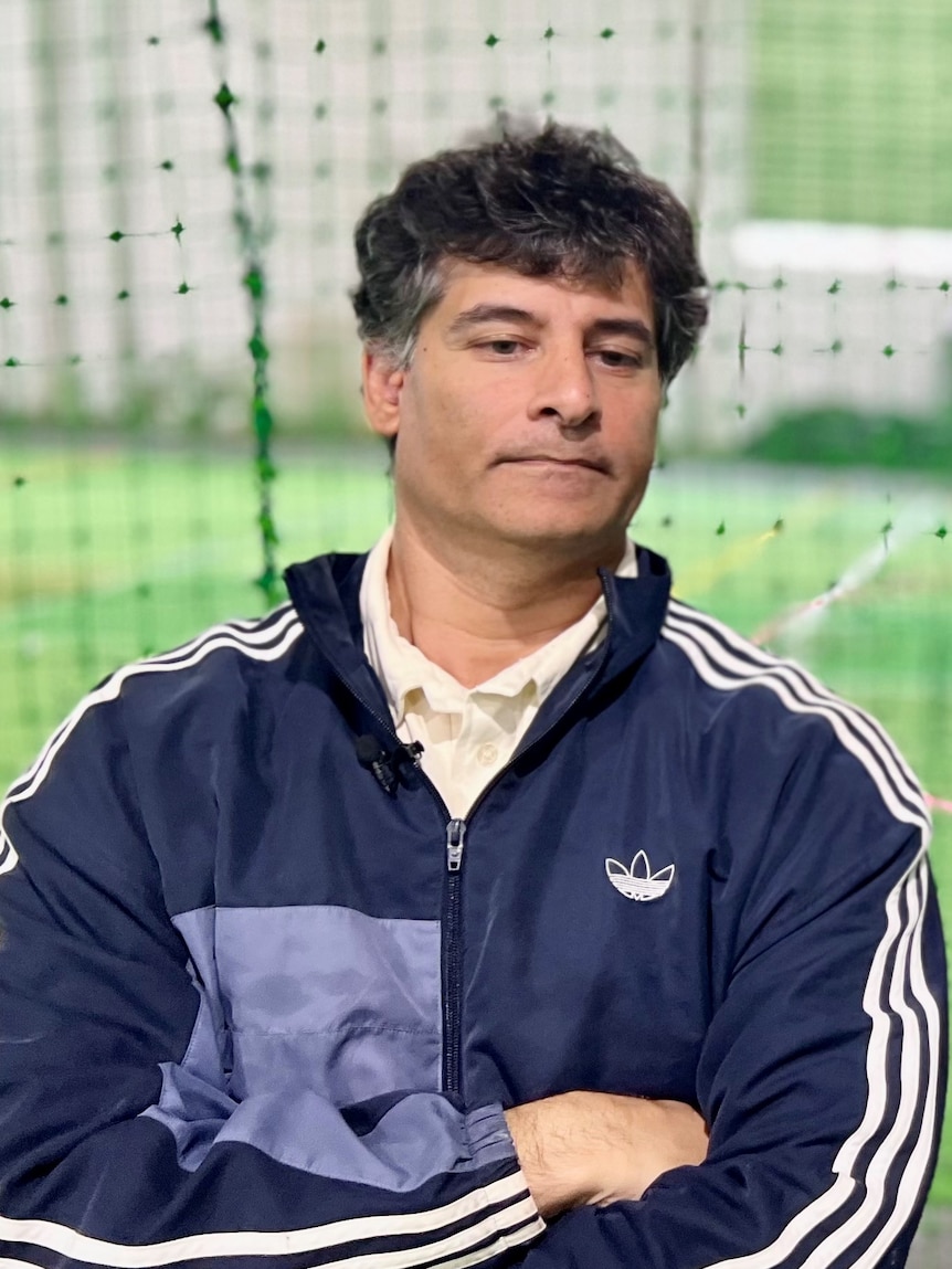 A man wearing a sports outfit leans in a fence with his arms crossed as a player holds up a cricket ball