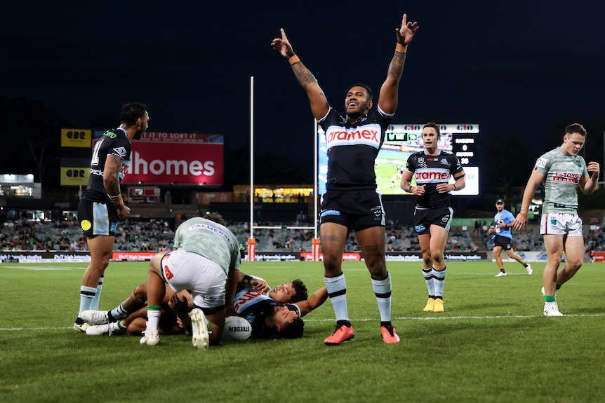 An NRL player holds his arms and index-finger in the air, celebrating, as a teammate scores next to him