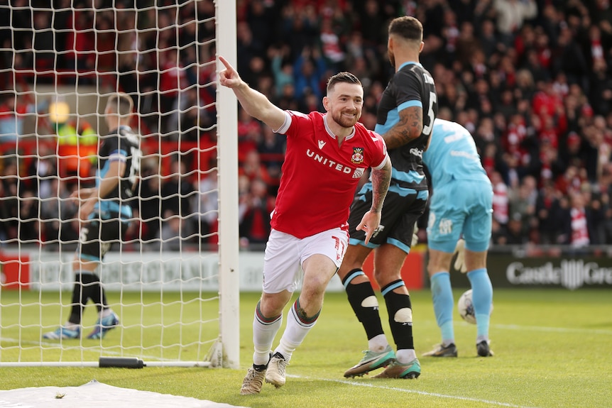 A Wrexham footballer runs away from goal smiling and pointing his finger in the air in celebration after a goal.