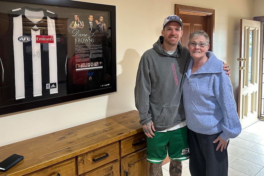 A man in a hoodie and shorts standing with his arm around an older woman standing next to a framed guernsey and plaque