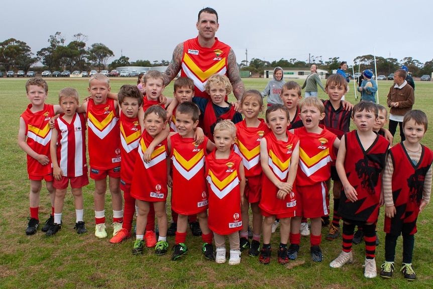 A group of kids dressed in red footy uniforms huddles around a man in the same uniform.