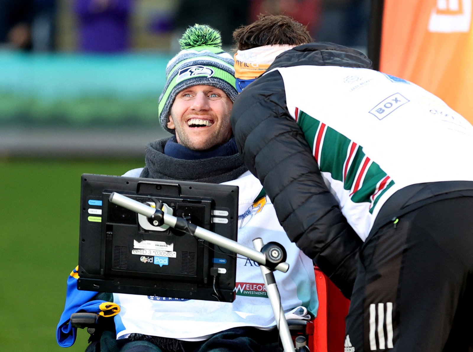 Rob Burrow smiles as he sits in a wheelchair and Kevin Sinfield speaks to him.