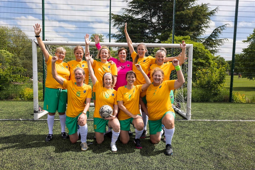 A group of older women in green and gold soccer get up kneeling in front of a soccer goal