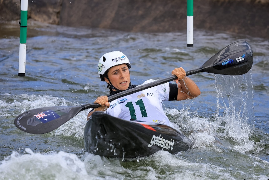 Noemie Fox competing in Canoe Slalom, holding the paddle across her chest, leaning back on the canoe to make it through a gate