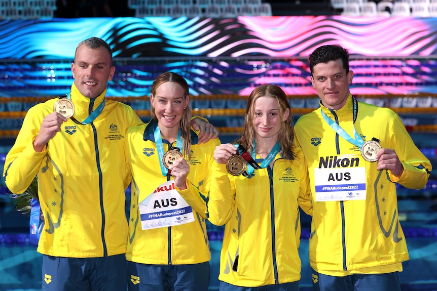 Swimmers wearing yellow jackets hold up medals and pose for a photo after winning a race.