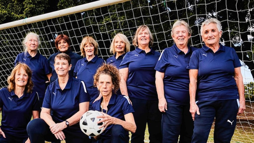 A group of older women wearing dark blue shirts pose for a photo holding a soccer ball in front of a goal