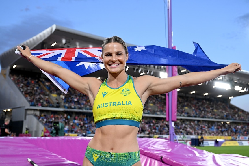 A beaming woman in an Australian athletics uniform holds an Australian flag behind her.