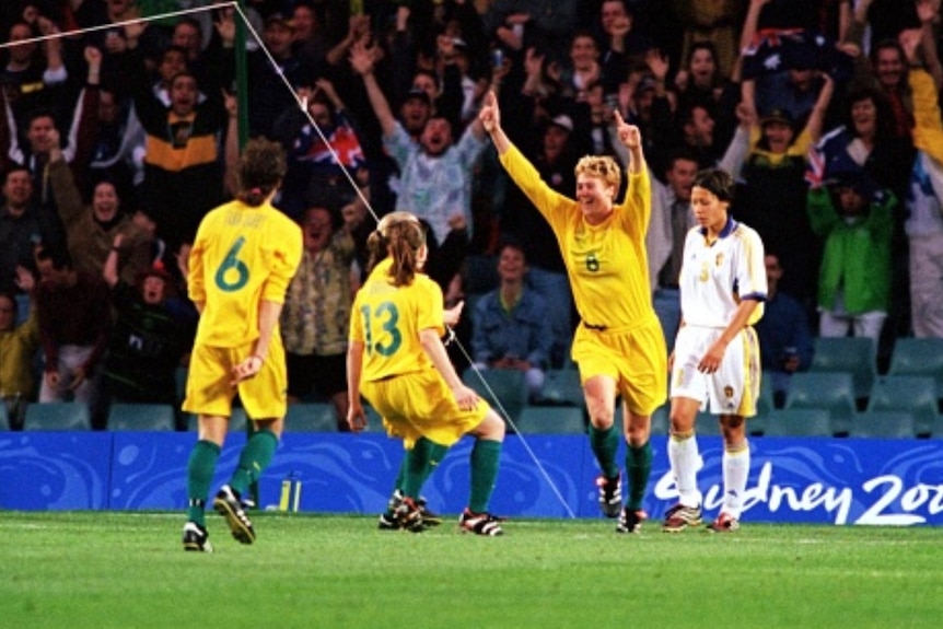 A women's soccer team wearing yellow and green celebrates after scoring a goal against a team in white in front of a big crowd