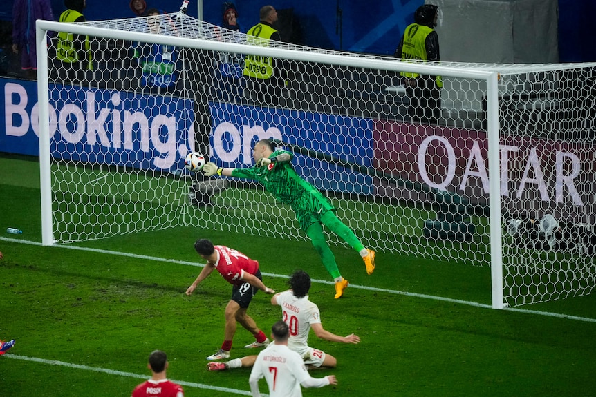 A goalkeeper dives across goal to get his fingertips to the ball to make a save as players look on.
