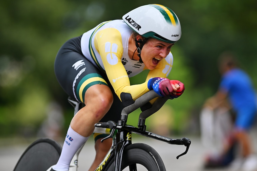 A female Australian road cyclist grimaces as she pedals fast while wearing an aerodynamic suit during a time trial stage.