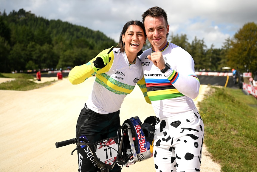 Two BMX riders, a woman wearing an Australian sporting outfit and a Frenchman wearing a rainbow top, pointing with a finger. 