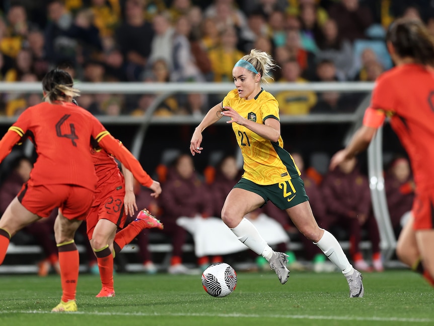 A Matildas footballer looks down at the ball as she runs, about to switch direction while a Chinese defender closes in.