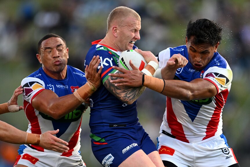 A NZ Warriors player grimaces as he holds the ball to his chest and is tackled by two Knights players.