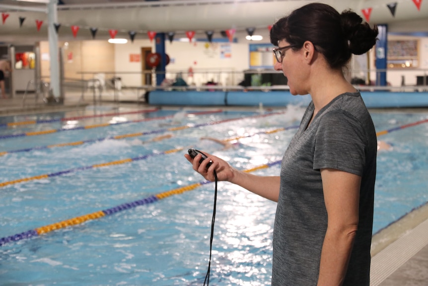 A woman holds a stop watch beside an indoor pool.