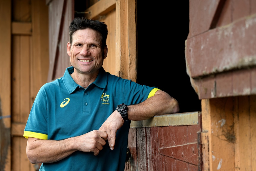 Equestrian Shane Rose leans on a horse stable and smiles at the camera.