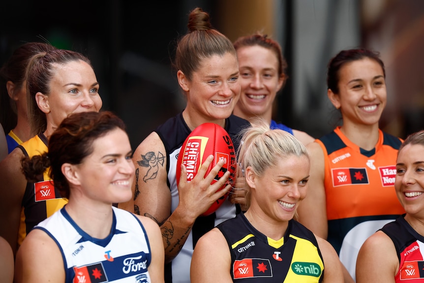 Bri Davey smiles while holding a football. She is surrounded by other club captains including Katie Brennan and Meg McDonald
