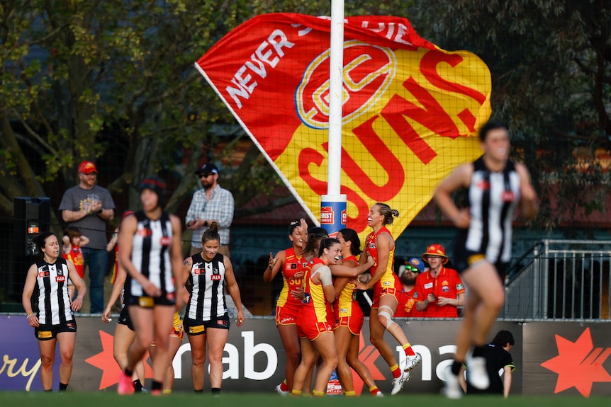 A group of Gold Coast Suns AFLW players embrace in the goalsquare after a goal, as fans wave a giant Suns flag behind the goals.