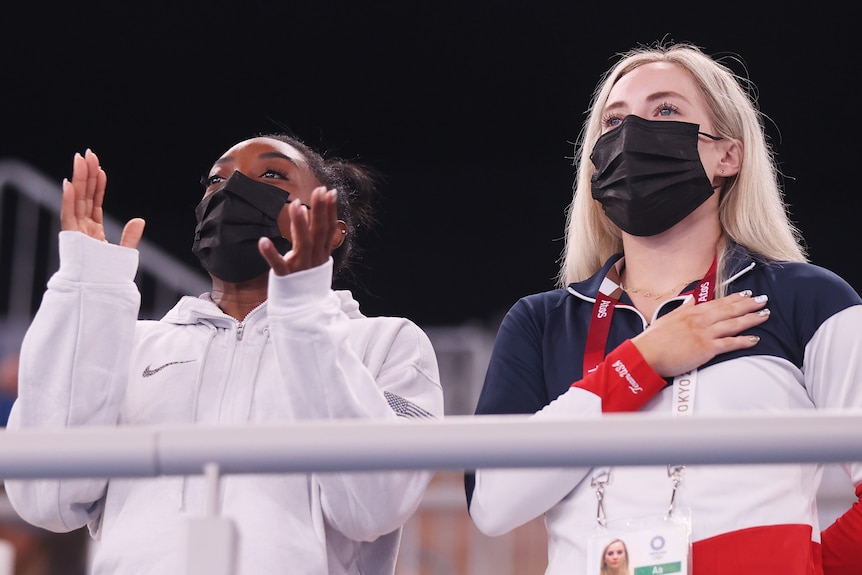 Two women wear masks at a sporting event, one claps the other has her hand on her heart.