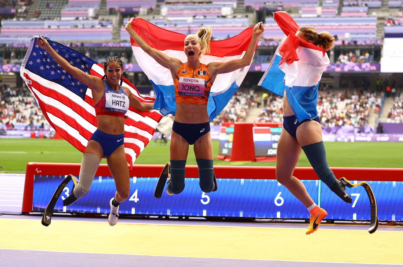 Three female long jumpers with prosthetic legs jump in the air, holding their country's flags behind their backs.