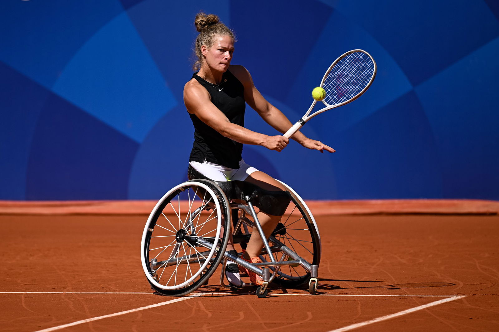 A woman in a wheelchair wearing a black singlet on a clay tennis court hits a tennis ball with a tennis racquet