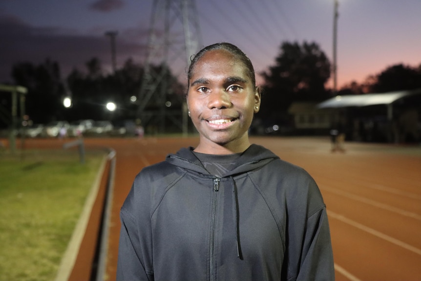 A young Aboriginal teen, Telaya, stares at the camera smiling. She wears a black jumper, the orange track behind her.