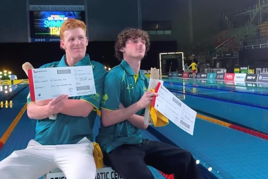 Two young men sit in front of indoor pool holding plane tickets