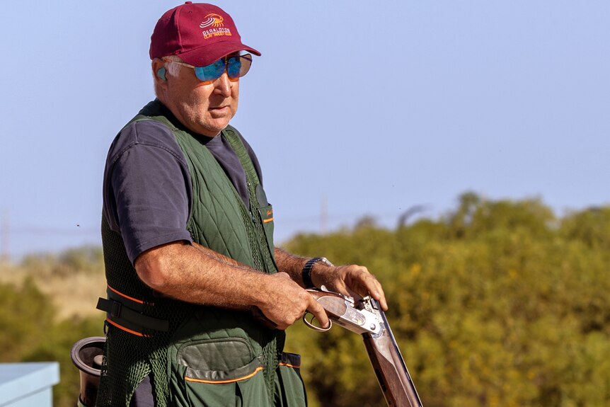 Man in red cap, sunglasses, and wearing ear plugs holds cracked shotgun as he places a new shell