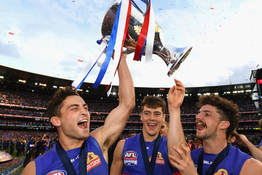 A group of three Western Bulldogs AFL players cheer with medals around their necks as one lifts the premiership cup.