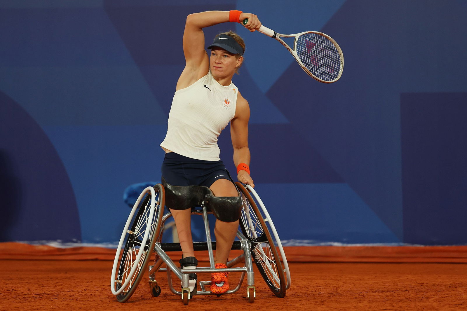 A female wheelchair user holds a tennis racquet over her head, like she's just hit a ball. She wears a white singlet and black shorts on a reddish brown clay tennis court and in front of a blue wall.