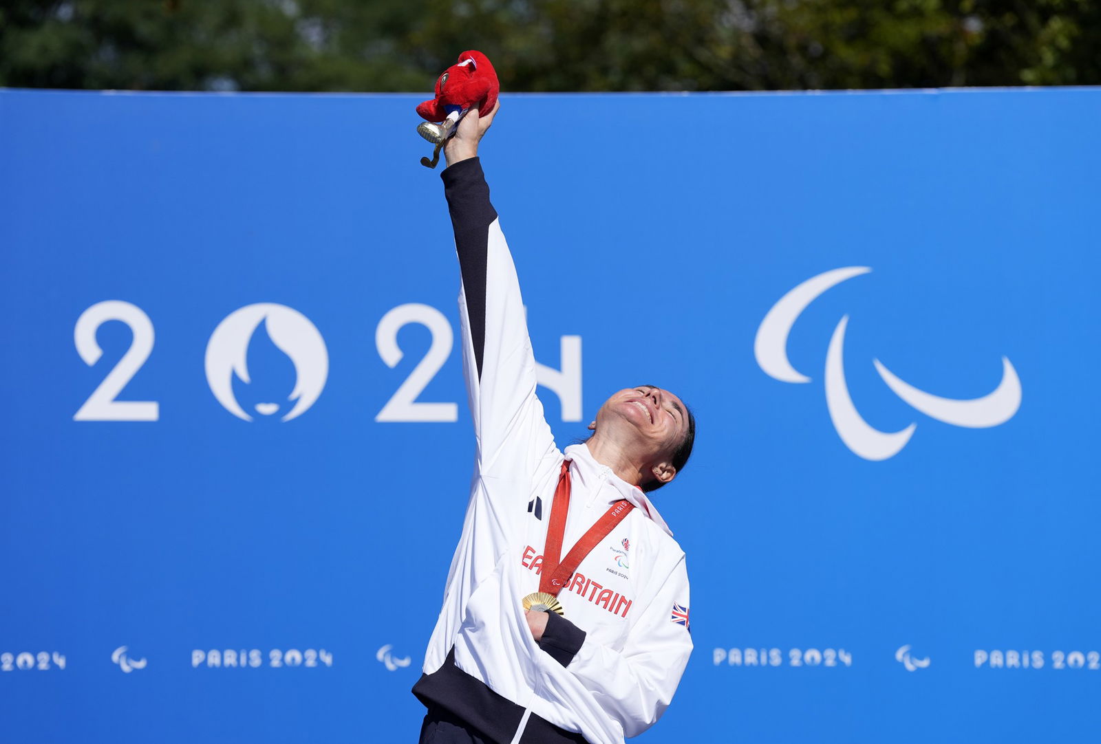 British Para cyclist Sarah Storey stands on top of the podium wearing her gold medal, with one arm raised in the air and her head thrown back with a broad smile.