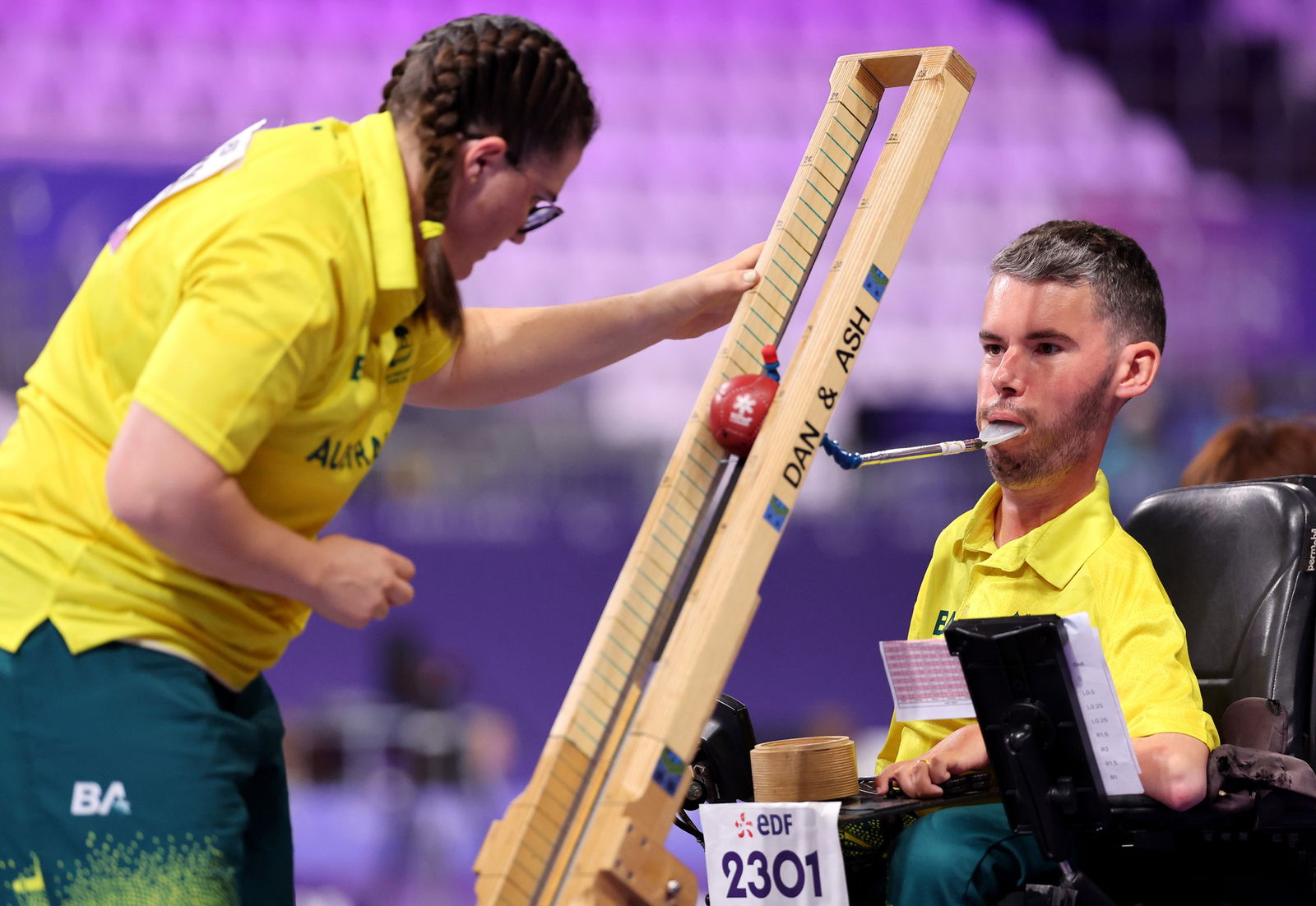 A man in a motorised wheelchair is propelling a red boccia ball down a wooden ramp with a device he is holding with his mouth. There is a woman in a yellow top holding the ramp steady and there are pink stadium seats behind them.