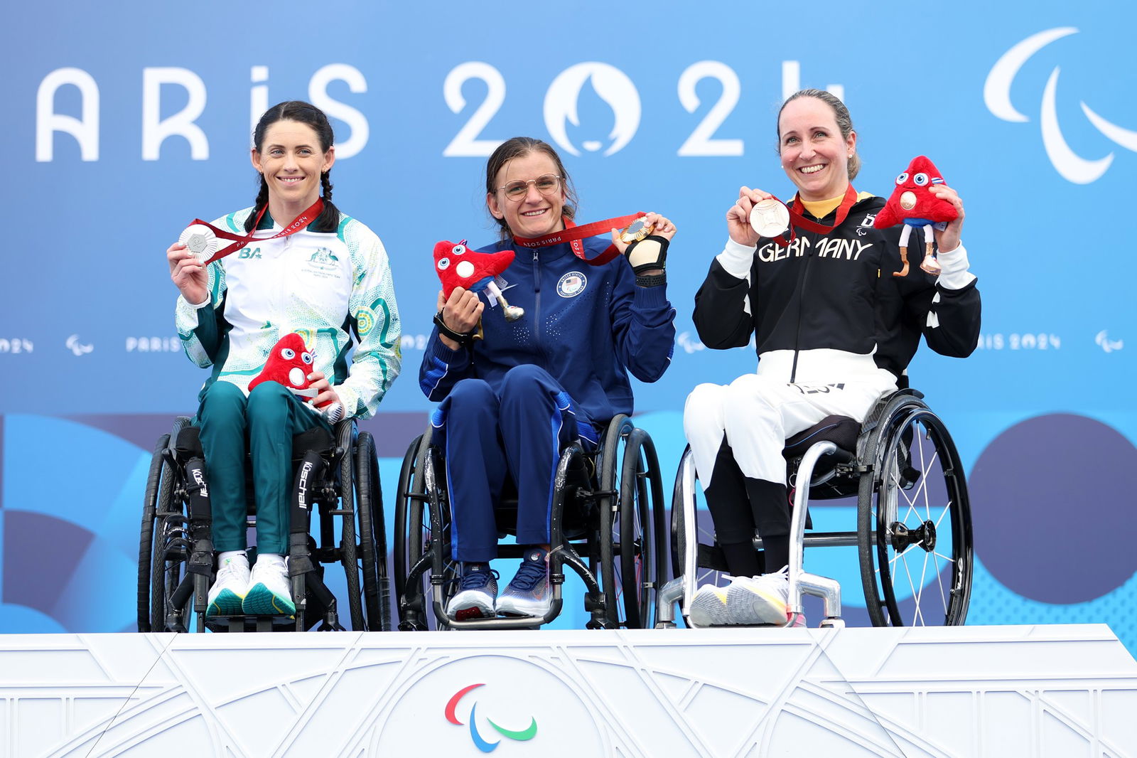 Three wheelchair para cyclists sit on the podium holding their medals and smiling.