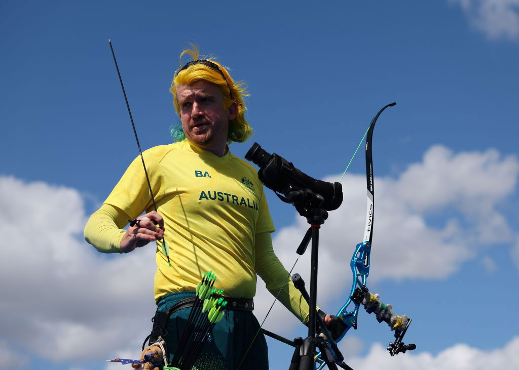 A man with a yellow shirt and yellow hair is holding an arrow in his right hand and a bow in his left. He has "Australia" written on his shirt. The background is a mostly blue sky with scattered clouds. It is a low angle shot.