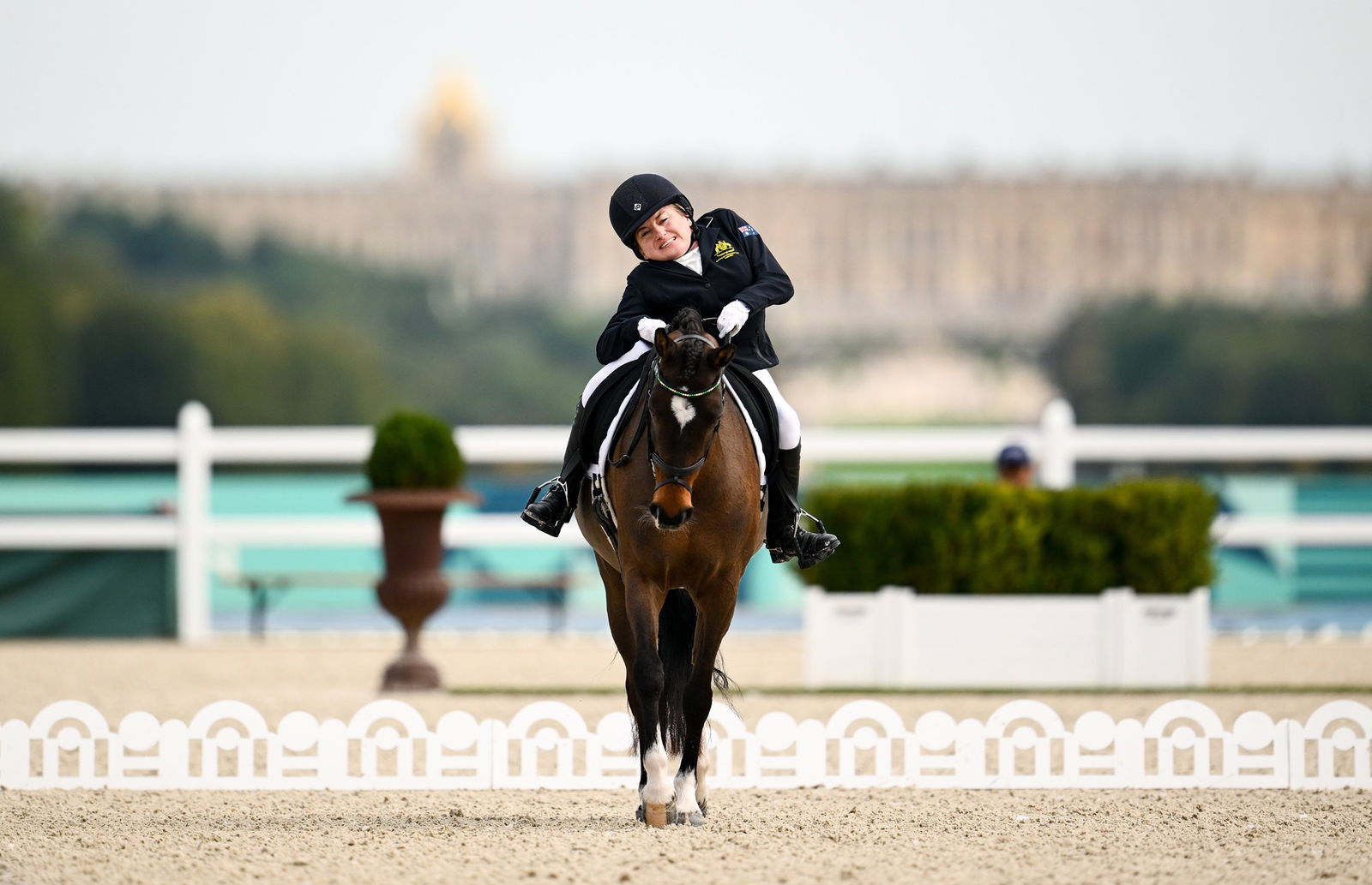 A mid shot of a woman riding a brown horse. She is wearing a black helmet and black riding gear, her body is leaning to her right. The horse is walking towards the photo taker on a brown, earthy floor with ornate decorations behind it. In the background, there is a fence and an ornate building.