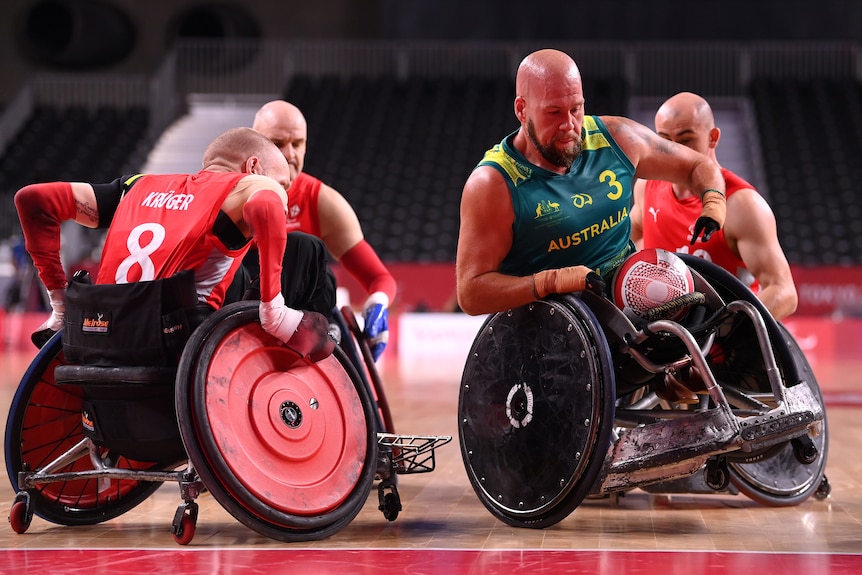 An Australian wheelchair rugby rolls his chair over the try line carrying the ball while surrounded by three defenders.