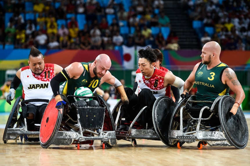 An Australian wheelchair rugby player carries the ball surrounded by two Japanese players as a teammate looks on.