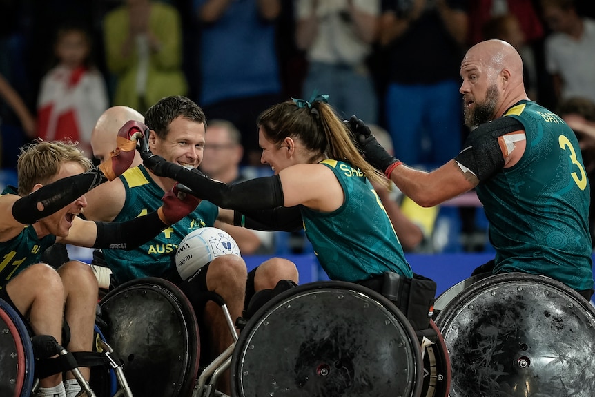 Three Australian wheelchair rugby players grin widely as they come together after a win, while a teammate sits behind.