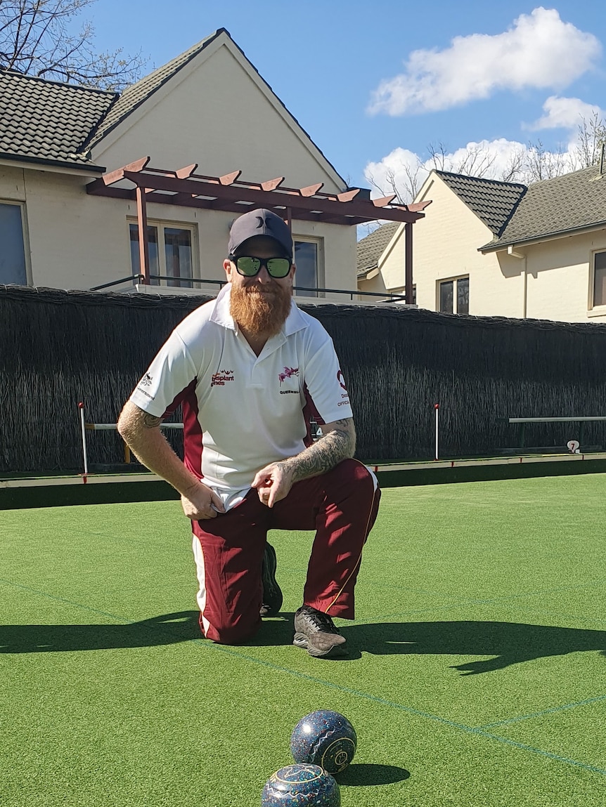 A man smiles, kneeling before some bowls on the green.