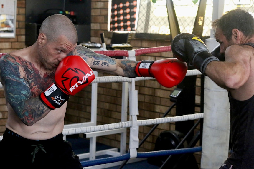 Man, buzz cut, tattoos on arm, chest, red glove, bare torso boxes with man with dark hair, black tee and gloves.