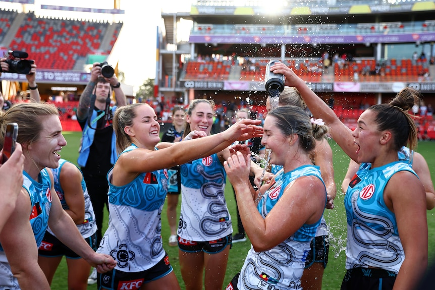 A group of Port Adelaide AFLW players celebrate on the ground after a win, pouring a drink over the head of a teammate.