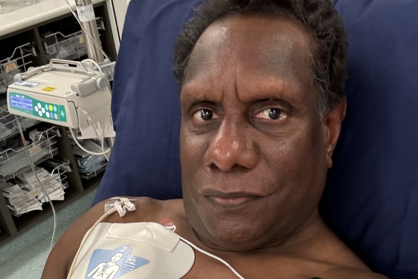 An unsmiling Indigenous man stares straight at a camera while in hospital, medical patches taped to his chest. 