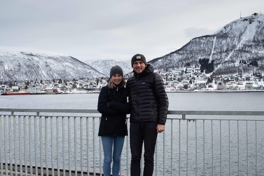 Reece and his wife wear black coats and post in front of the water, with snowy mountains visible in the background.