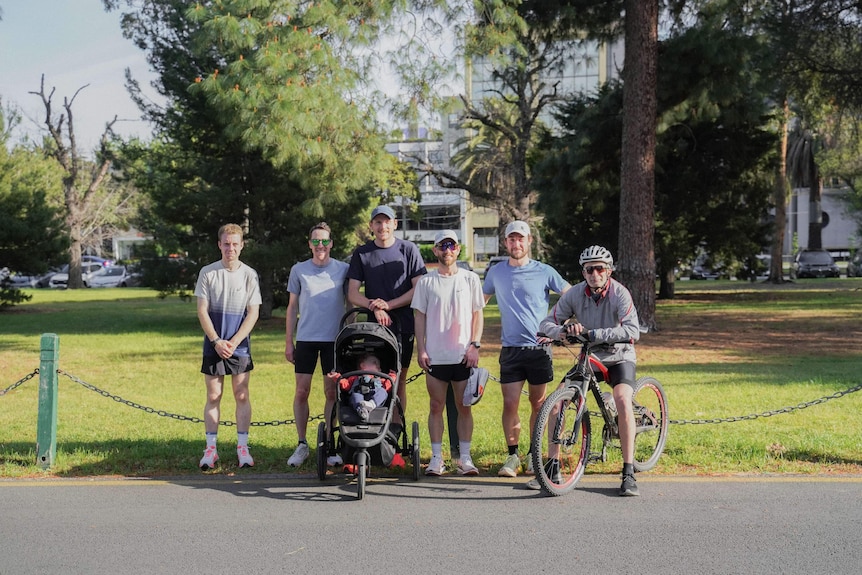 Reece Edwards, wearing runnign gear and leaning on a pram with a baby inside, in a group photo at a park.