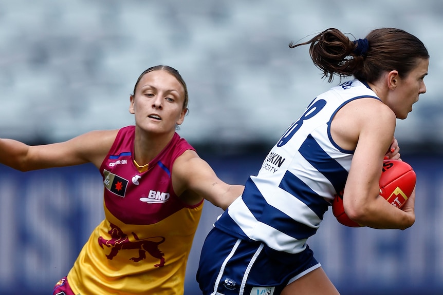 A Geelong AFLW player runs hard carrying the ball as a Brisbane player tries to tackle her.