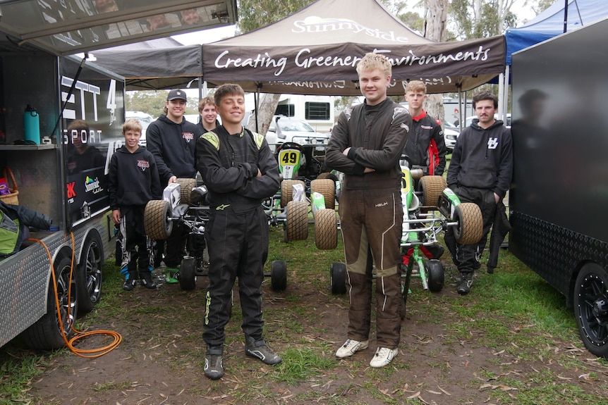 Two teenage boys wearing black coveralls with others behind between two black trailers