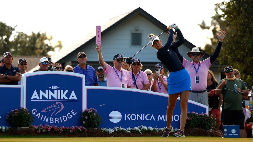 American golfer Nelly Korda completes her swing on a tee shot as marshals hold up "quiet signs" and the crowd watches.
