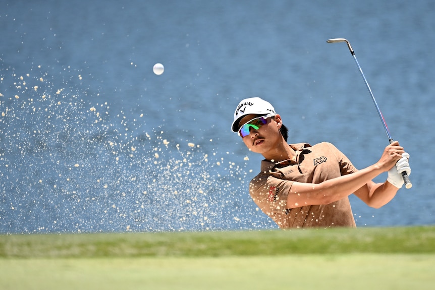 Golfer follows through after a shot out of the bunker, with sand flying in the air
