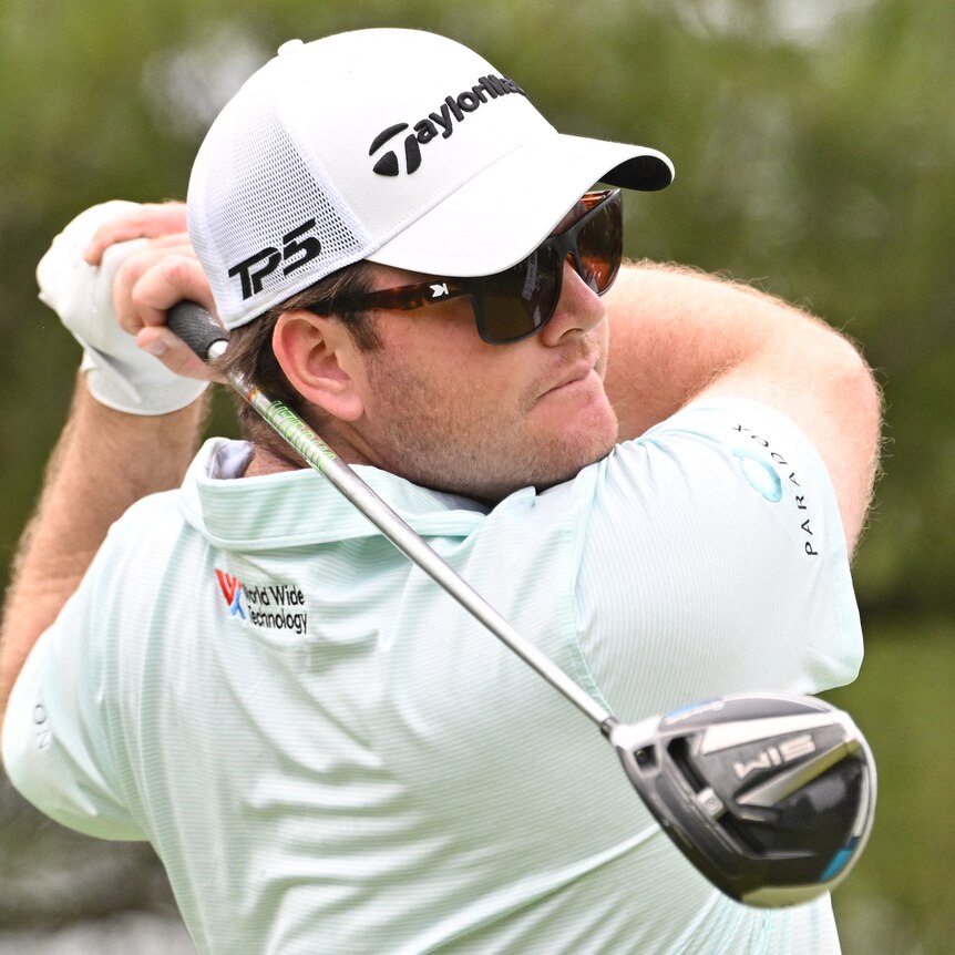 Harry Higgs hits his tee shot, wearing sunglasses and a white cap
