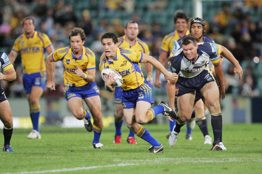 A man in a sports uniform holds a ball and runs with his mouth open while being chased by other players.
